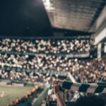 Es ist die Stadionkurve bei einem Abendspiel zu sehen. Das Foto wurde aus der Kurve geschossen und alle Fans halten Handylichter oder Schals hoch.