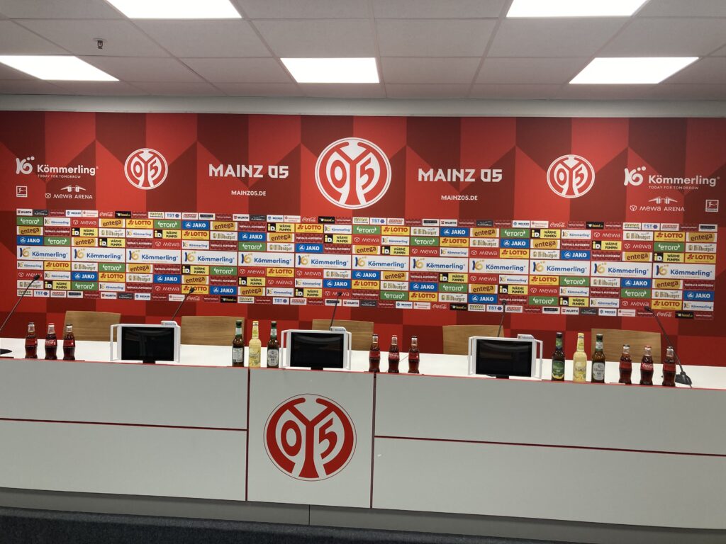 Hier sieht man den Presseraum von Mainz 05 mit der Sponsorenwand im Hintergrund und dem Tisch an dem Trainer und Spieler nach dem Spiel Journalisten Rede und Antwort stehen. Es sind 5 Sitzplätze und 5 Mikrofone zu sehen.