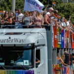 Das Bild zeigt in der linken unteren Ecke einen grauen LKW. Auf dem Aufleger des LKWs ist ein zweistöckiges Podest aufgebaut. Auf dem Podest stehen ganz viele Menschen eng an eng. Sie halten Flaggen in der Hand, lehnen sich vom LKW runter und werfen Dinge. Auch um den LKW herum ist eine große, bunte Menschenmenge zu erkennen.