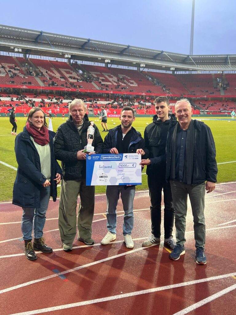 Auf dem Bild sind fünf Menschen auf der Laufbahn in einem Stadion zu erkennen. IN der Mitte stehen Lucas und Maxim, zwei Mitarbeiter von T_OHR. Links und rechts neben ihnen stehen Hort Hrubesch und Christian Streich sowie eine dritte Offizielle. Lucas und MNAxim Halen einen übergroßen weißen Scheck in der Hand. Auf diesem Scheck ist ein Preisgeld von 5.000 Euro festgeschrieben.