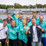 Auf dem Bild erkennt man eine Gruppe mit 8 jungen Menschen vor einem Fluss. Sie alle lächeln und jubeln in die Kamera. Viele von ihnen tragen die Uniform der "Rhein Ruhr 2025 FISU World University Games".