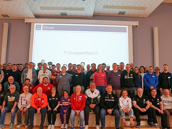Das Foto zeigt die rund 70 Gäste der Expert*innenforums 2026. In drei Reihen sind sie vor einer Leinwand aufgestellt, auf der in blauer Schrift auf weißem Hintergrund das Wort "Gruppenfoto" zu lesen ist. Sie sind in verschiedenen Farben gekleidet, einige Tragen Jacken und T-Shirts von ihren Fußballvereinen.