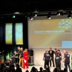 Das Bild zeigt die Bühne in der Tafelhalle in Nürnberg. Auf der Bühne ist im Hintergrund eine große Leinwand zu erkennen, auf dem das abstrakte Logo des Deutschen Fußball-Kulturpreises zu erkennen ist. Vor der Leinwand stehen einige Menschen auf der Bühne. Darunter Sportmoderatorin Kathrin Müller-Hohenstein, Jürgen Klopp und einige der Preisträger des Abends. Sie alle tragen schicke Abendgarderobe und stellen sich gemeinsam für ein Foto auf.