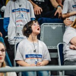 Das Foto zeigt ein Kind, das mit Kopfhörern und einem Empfängergerät auf den Zuschauerrängen der SAP Arena sitzt. Staunend schaut es nach oben. Es trägt ein graues T-Shirt, auf dem das Logo der Spardabank zu erkennen ist.