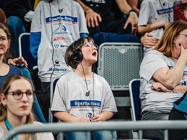 Das Foto zeigt ein Kind, das mit Kopfhörern und einem Empfängergerät auf den Zuschauerrängen der SAP Arena sitzt. Staunend schaut es nach oben. Es trägt ein graues T-Shirt, auf dem das Logo der Spardabank zu erkennen ist.