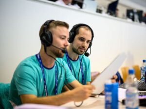 Von links vorne sieht man in die Blindenreporter Lucas Schmelz und Luis Bracht mit Headsets, die auf einer Medientribüne sitzen. Sie schauen auf ein Blatt Papier, das Lucas in der Hand hält.