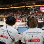 Von hinten sieht man die Reporter*innen Louis (links) und Ann-Marit (rechts) direkt am Basketballfeld sitzend. Sie tragen beide ein Headset und ein weißes T-Shirt mit der Aufschrift "Blindenreportage / Live-Audiodeskription" und dem Logo von T_OHR. Im Hintergrund sieht man leicht unscharf das Basketballfeld, auf dem eine deutsche Spielerin (in schwarz), eine spanische Spielerin (in weiß) und ein Schiedsrichter (in grau) stehen.
