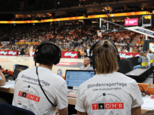 Von hinten sieht man die Reporter*innen Louis (links) und Ann-Marit (rechts) direkt am Basketballfeld sitzend. Sie tragen beide ein Headset und ein weißes T-Shirt mit der Aufschrift "Blindenreportage / Live-Audiodeskription" und dem Logo von T_OHR. Im Hintergrund sieht man leicht unscharf das Basketballfeld, auf dem eine deutsche Spielerin (in schwarz), eine spanische Spielerin (in weiß) und ein Schiedsrichter (in grau) stehen.