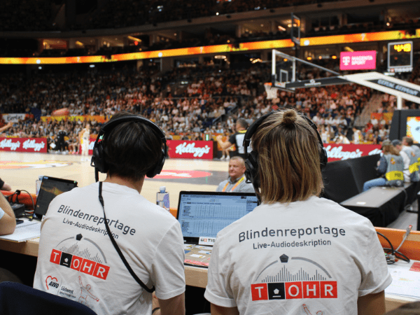 Von hinten sieht man die Reporter*innen Louis (links) und Ann-Marit (rechts) direkt am Basketballfeld sitzend. Sie tragen beide ein Headset und ein weißes T-Shirt mit der Aufschrift "Blindenreportage / Live-Audiodeskription" und dem Logo von T_OHR. Im Hintergrund sieht man leicht unscharf das Basketballfeld, auf dem eine deutsche Spielerin (in schwarz), eine spanische Spielerin (in weiß) und ein Schiedsrichter (in grau) stehen.